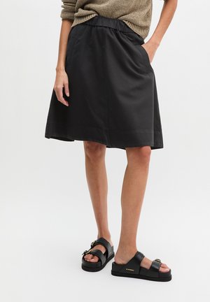 Vrouw die een zwarte rok tot op de knie draagt met een elastische taille en zwarte sandalen met gesp, staand met één hand in de zak.