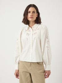 Witte blouse met opgezette lange mouwen, met bloem borduurwerk op de mouwen. Knopsluiting met een getailleerde kraag, gecombineerd met beige broeken.