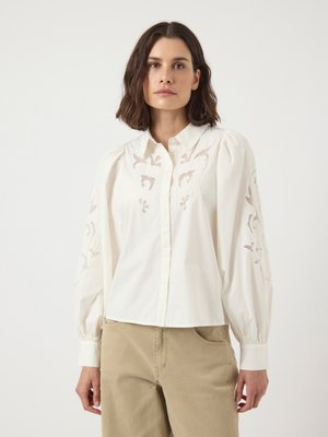 White blouse with puffed long sleeves, featuring floral embroidery on sleeves. Button-down design with a tailored collar, paired with beige pants.