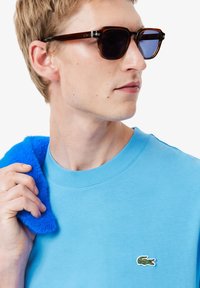 T-shirt di cotone blu con collo rotondo, caratterizzata da un piccolo logo di coccodrillo verde. Il modello tiene un oggetto blu e peloso sulla spalla mentre indossa occhiali da sole.