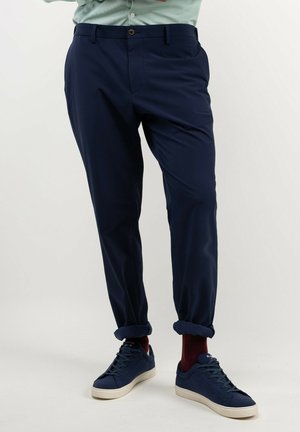 Hombre con pantalones azul marino remangados en los puños, calcetines rojo oscuro y zapatillas azul marino con suelas blancas, de pie contra un fondo liso.