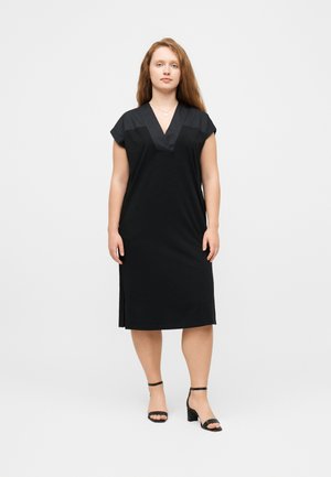 Femeie cu păr lung roșcat, purtând o rochie midi neagră cu decolteu în V și sandale negre cu toc, stând în picioare pe un fundal simplu și deschis la culoare.