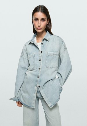 Femme portant une veste en denim bleu clair surdimensionnée et un jean assorti, se tenant avec une main dans la poche devant un fond uni.