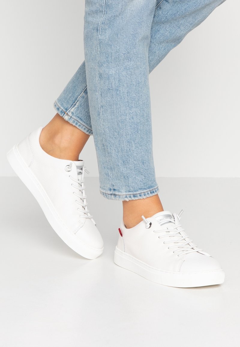 Baskets en cuir blanc avec bout rond, design à lacets et semelle extérieure plate en caoutchouc blanc. Caractérisées par un accent rouge sur le talon et une texture subtile.