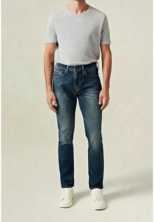 Homme portant un t-shirt gris clair à manches courtes, un jean bleu foncé avec des détails délavés et des baskets blanches, debout sur un fond uni.