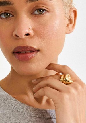 Kuldne skulpturaalne sõrmus, mis on kantud hele naha tooniga käel, sileda tekstuuri ja pehme läikega. Peal nähtav halli särgi kaelus.