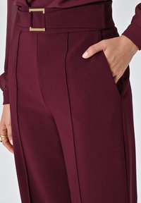 Pantalons bordeaux taille haute avec une jambe large, présentant des plis devant, des poches latérales et une ceinture accentuée par une boucle carrée en or. Texture de tissu lisse.