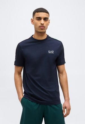 Fiatal férfi rövid hajjal, sötétkék EA7 Emporio Armani pólót visel, vállán piros, fehér és zöld csíkokkal, valamint sötétzöld nadrágot.