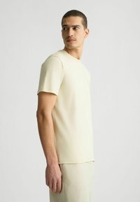 Hellbeige Kurzarm-T-Shirt aus einem glatten, weichen Stoff, mit einem klassischen Rundhalsausschnitt und geradem Saum, ergänzt durch ein minimales Design.