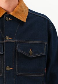 Donkerblauwe spijkerjack met een corduroy kraag, voorzien van messing knopen en oranje stiksels, inclusief twee voorzakken en een logo detail.