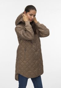 Ung kvinna justerar huven på en brun vadderad jacka med dragkedjeficka, iförd blå jeans mot vit bakgrund.