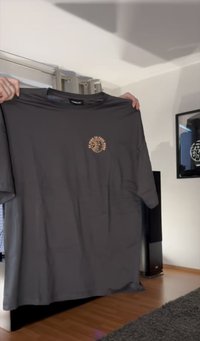 Dunkelgraues T-Shirt mit einer kleinen kreisförmigen Pfirsichgrafik und Text auf der linken Brust, von zwei Händen drinnen gehalten.