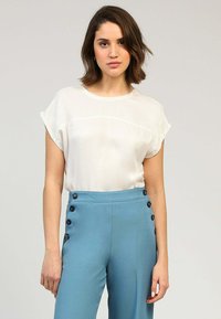 Chemisier blanc à manches courtes avec un col rond, associé à un pantalon bleu taille haute orné de boutons décoratifs noirs sur le côté.