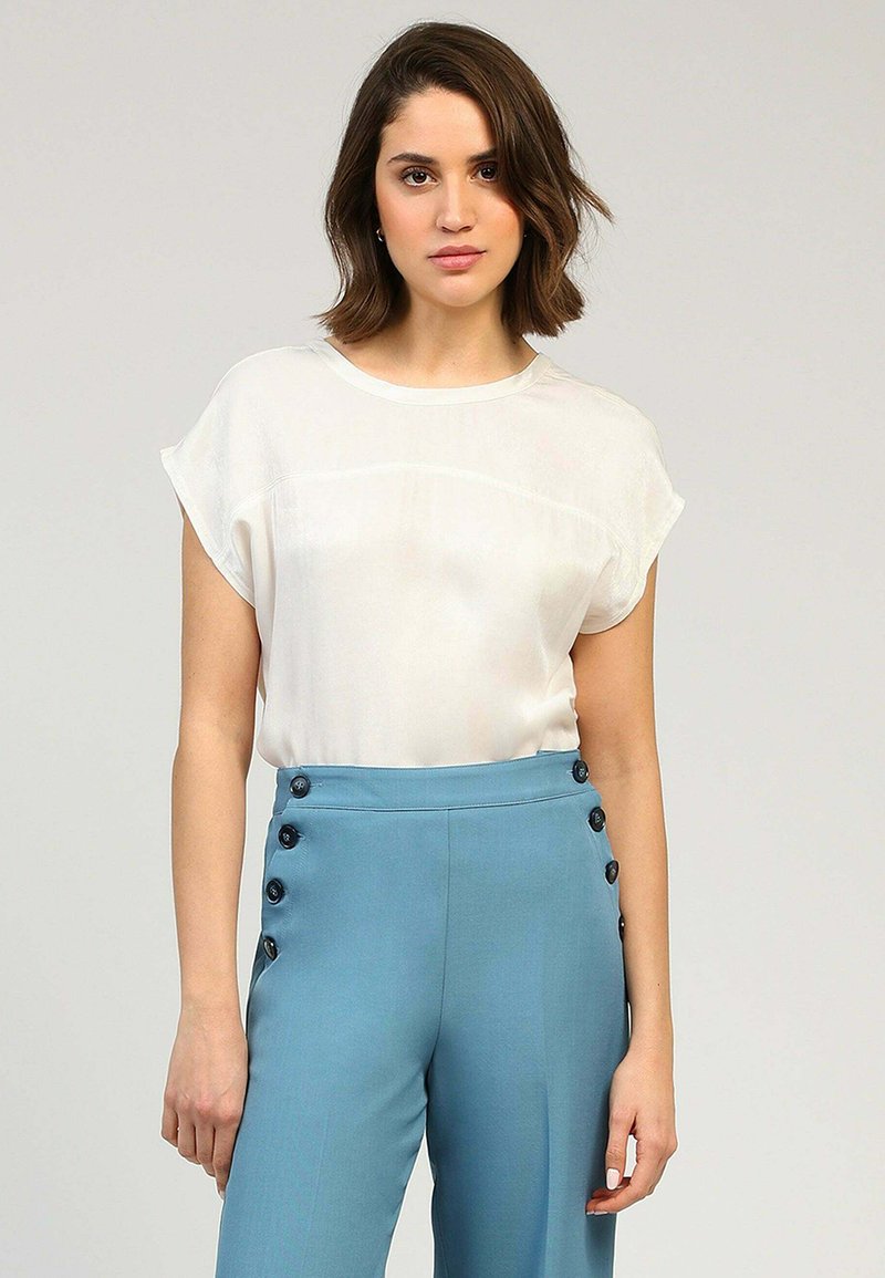 Chemisier blanc à manches courtes avec un col rond, associé à un pantalon bleu taille haute orné de boutons décoratifs noirs sur le côté.