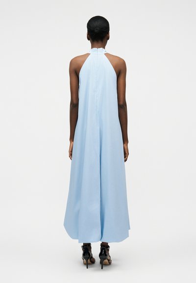 Femme à la peau foncée et aux cheveux attachés en chignon, vêtue d'une robe sans manches à col halter bleu clair et de sandales à talons noirs, debout de dos.