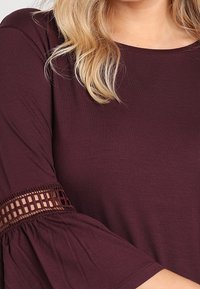 Viininpunainen yläosa, jossa liehuvat hihat ja kalaverkkokuvioiset leikkaukset hihansuiden kohdalla. Valmistettu pehmeästä ja sileäpintaisesta kankaasta.