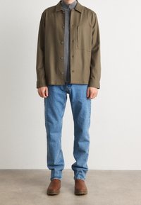 Chaqueta de botones de color verde oliva con un bolsillo en el pecho sobre una camiseta gris con cremallera, combinada con jeans de color azul claro y botas de tobillo marrones.