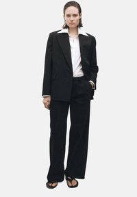 Črn dvojniki blazer z belim ovratnikom, v kombinaciji z ohlapnimi črnimi hlačami in črnimi sandalami. Gladka tkanina in krojen dizajn.