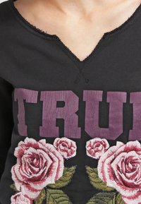 Černé bavlněné tričko s výstřihem do V, zdobené embosovaným fialovým nápisem "TRUE" a detailní vyšívanou růží v růžové barvě se zelenými listy.