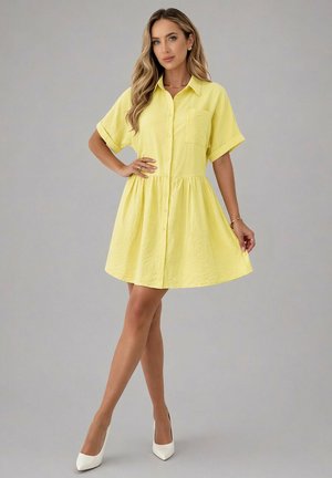 Femme portant une robe jaune à manches courtes, boutonnée avec col et talons blancs, posant avec une main sur la taille et l'autre tenant l'ourlet de la robe.