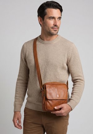 Mężczyzna noszący beżowy sweter i brązowe spodnie, trzymający brązową skórzaną torbę na ramię z klapką i zamkiem błyskawicznym.