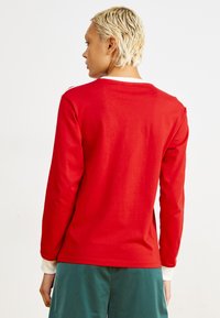 Rød langermet topp med rund hals, ensfarget tekstur og hvit detalj ved mansjettene. Båret med grønne shorts, sett fra baksiden.