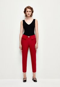 Pantalones rojos ajustados con bolsillos frontales y detalles en dorado, combinados con una blusa negra sin mangas. La tela parece suave y ajustada.