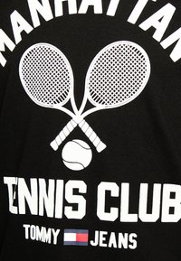 Juodas marškinėliai su baltu grafiniu dizainu, vaizduojančiu kryžmines teniso raketes ir teniso kamuoliuką, taip pat su tekstu "MANHATTAN TENNIS CLUB".
