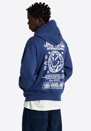 Persoon die een blauwe hoodie draagt met witte grafische tekst, symbolen en een glimlachende globe op de achterkant, gecombineerd met een donkere broek en een wit overhemd.