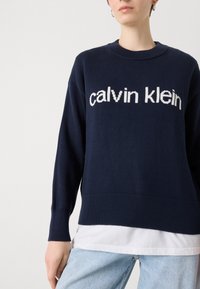 Tamsiai mėlynas megztinis su pynimu apatine dalimi ir rankovėmis, su baltu užrašu "calvin klein", išryškintu priekyje.
