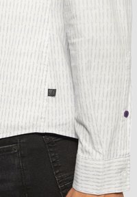 Witte shirt met smalle paarse verticale strepen. Het heeft een knoopmanchet met een paarse knoop en een klein zwart label op de zijnaad.