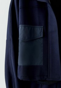 Marineblauwe jas van zacht materiaal met een contrasterende matte zak. De zak heeft een klep en is met rechte lijnen gestikt.