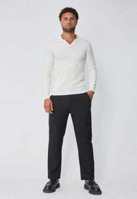 Pull blanc en maille avec un col en V et un col, associé à un pantalon noir fuselé et des chaussures noires, mettant en valeur un tissu texturé.