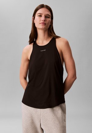 Kvinne med langt brunt hår iført en svart ermeløs Calvin Klein-topp og lysegrå joggebukser, stående mot en ensfarget grå bakgrunn.