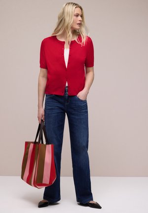 Femme aux longs cheveux blonds, portant un haut rouge à manches courtes boutonné, un jean bleu, des ballerines noires, tenant un sac fourre-tout rayé multicolore.