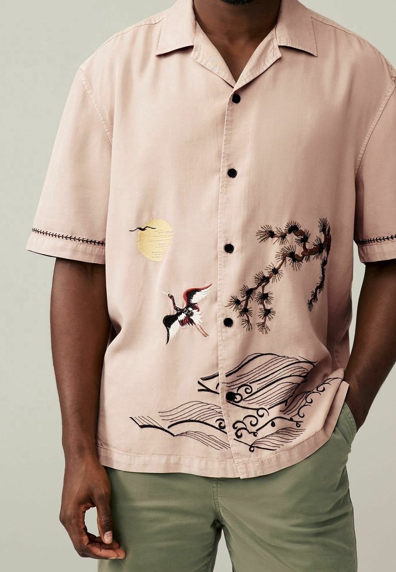 Koszula z krótkim rękawem w jasnoróżowym kolorze z czarnymi guzikami, z misternym haftem przedstawiającym żurawia, gałęzie sosny i słońce z przodu.
