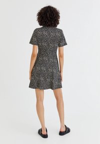 Schwarzes Kurzarmkleid mit weißem Blumenmuster, ausgestelltem Rock und weichem Stoff. Getragen mit schwarzen Slip-On Sandalen. Rückansicht.