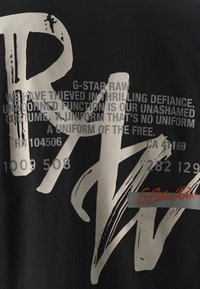 Camiseta negra con un gran texto gráfico en blanco, que presenta letras en negrita y detalles impresos pequeños. Incluye el nombre de la marca "G-Star Raw" en naranja.