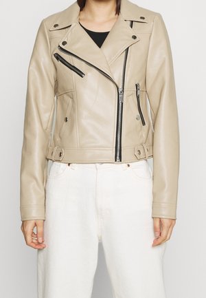 Veste biker courte en cuir beige avec des fermetures éclair noires et des boutons-pression, portée sur un haut noir et assortie à un pantalon blanc.