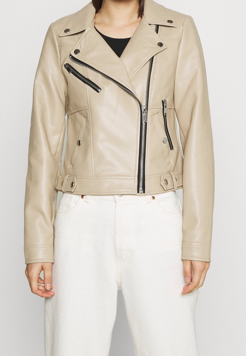 Chaqueta de cuero beige recortada estilo biker con cremalleras negras y botones de presión, usada sobre una camiseta negra y combinada con pantalones blancos.