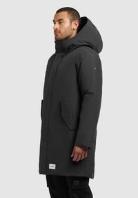 Zwarte waterdichte parka met een capuchon, voorzien van twee zijzakken, een ritsluiting en een logo-embleem nabij de zoom. Glad van textuur.