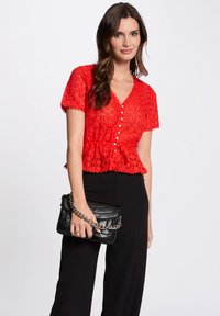Blouse en dentelle rouge à manches courtes, bords festonnés, fermeture à boutons sur le devant et taille peplum. Associe à un pantalon large noir et un sac à main noir.
