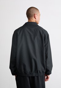 Black windbreaker jacket with a relaxed fit, elastic hem, and a collar. Smooth texture, features minimalistic design without visible branding.