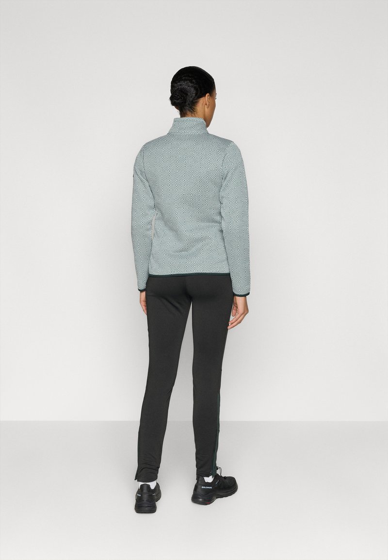 Jasnoniebieska kurtka polarowa w diamentowy wzór, z wysokim kołnierzem i czarnymi wykończeniami, zestawiona z czarnymi spodniami slim fit i czarnymi butami sportowymi.