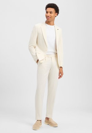 Jeune homme souriant, portant un costume sur mesure écru avec une chemise blanche et des mocassins beige, debout avec une main dans la poche.