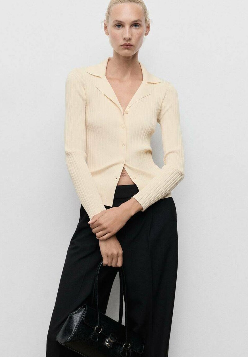 Cardigan a costine crema con maniche lunghe e chiusura a bottoni, abbinato a pantaloni neri larghi e a una piccola borsa nera strutturata.