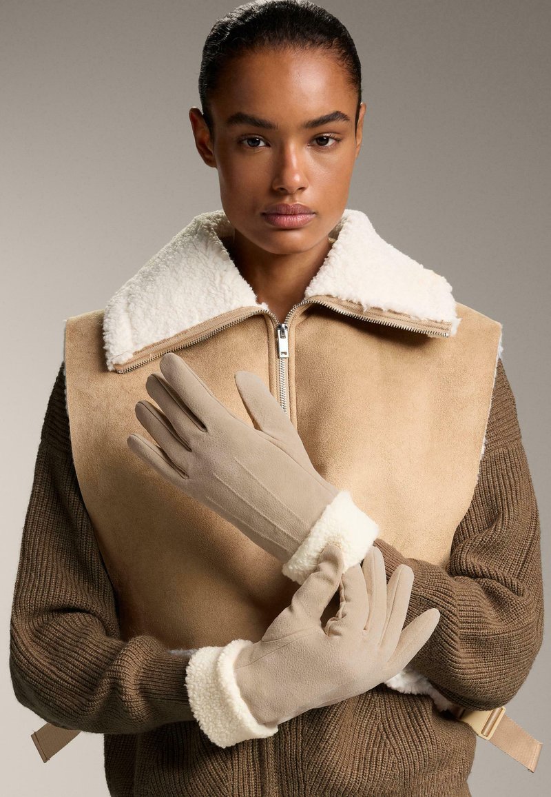 Beige rukavice s měkkým, plyšovým bílým lemem, doplněné hnědým žebrovaným svetrem a béžovou vestou s ovčí vlnou.