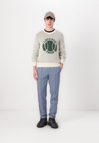 Pulover gri deschis cu logo-ul „LACOSTE PARIS” verde, tricot texturat. Pantaloni albaștri cu model și dungi laterale. Adidași negri. Șapcă bej.