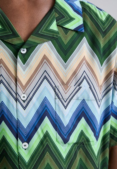 Chemise à manches courtes avec un motif zigzag multicolore en vert, bleu et beige. Tissu doux, fermeture à boutons et détail de poche poitrine.