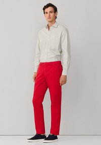 Uomo in piedi che indossa una camicia a righe bianche e grigie con bottoni, pantaloni rossi vivaci e scarpe da ginnastica nere, su uno sfondo grigio chiaro uniforme.